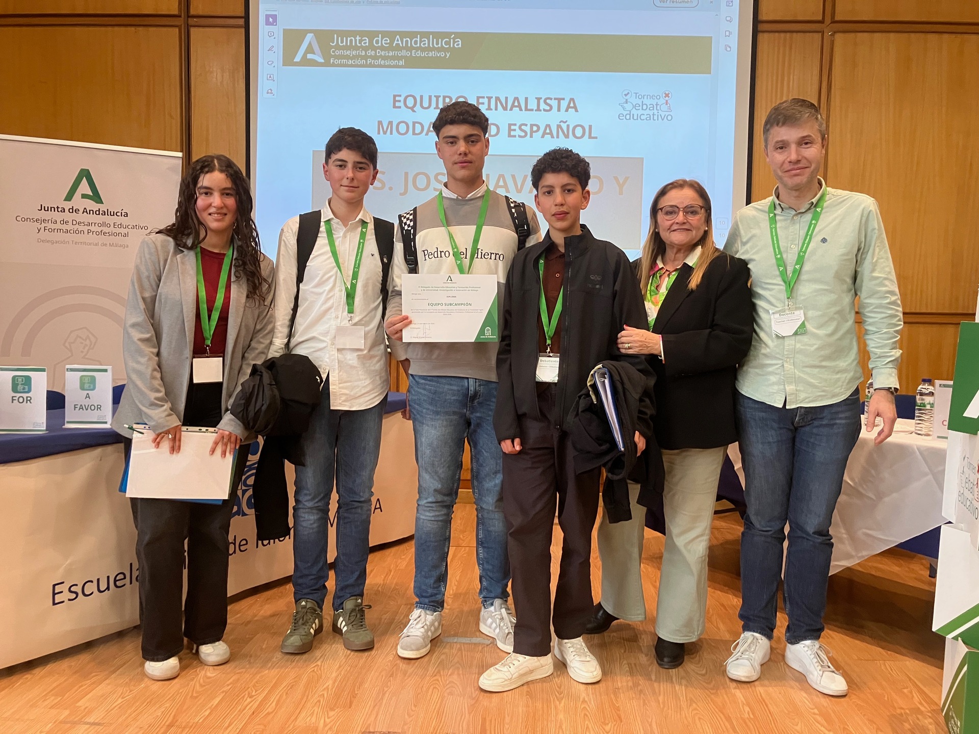 Seis personas posan en un evento educativo, con credenciales verdes y un diploma en el centro. Torneo de debate Málaga. IES José Navarro y Alba.